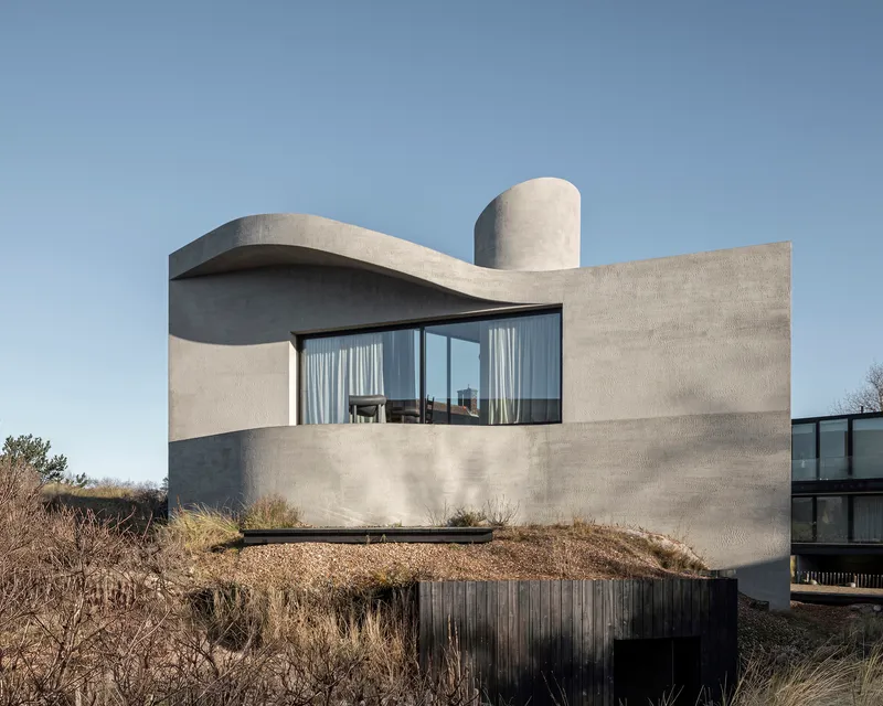 Cast concrete facade with curved roof element and cylindrical volume atop a dune landscape