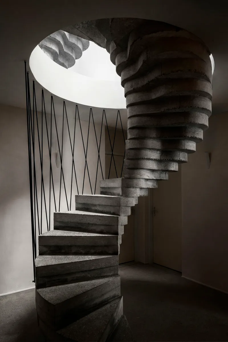 View up the spiral concrete staircase with zigzag steel balustrade beneath a circular skylight