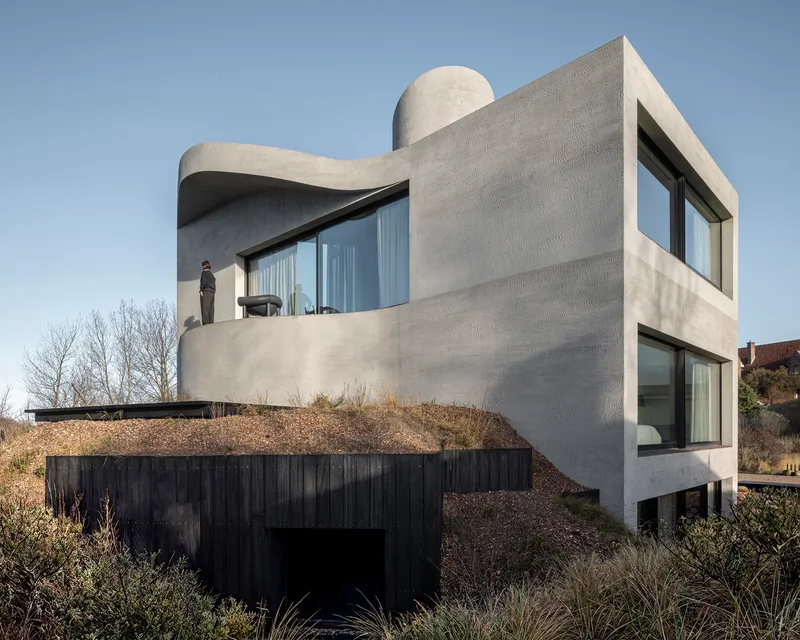 Curved concrete volume with recessed terrace and figure standing beside glazed doors overlooking dune grasses