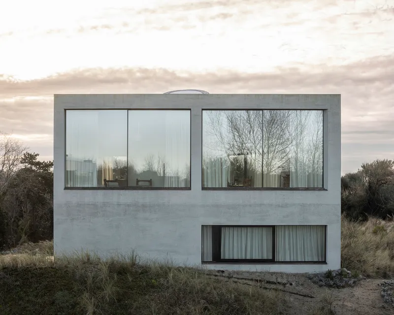 Rear facade with paired horizontal windows overlooking coastal dunes under overcast sky