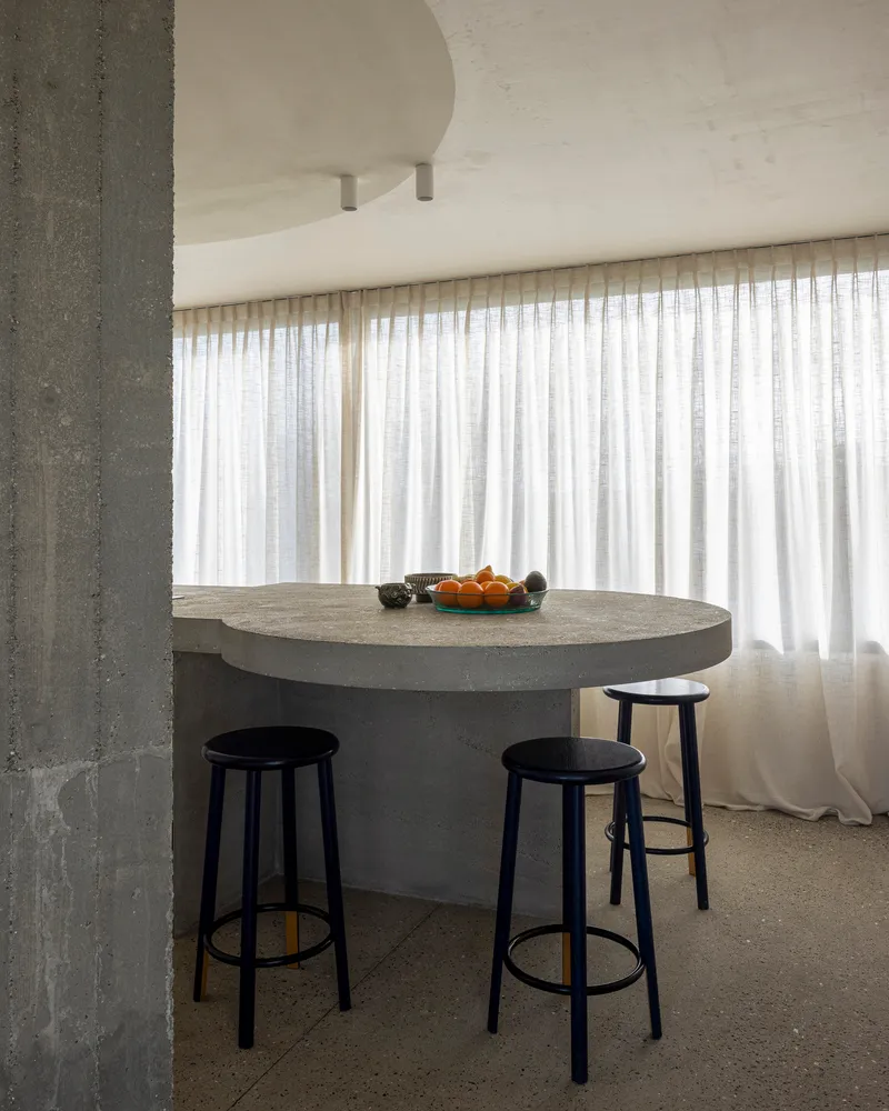 Curved concrete breakfast counter with black stools beneath sheer curtains and circular ceiling pendant