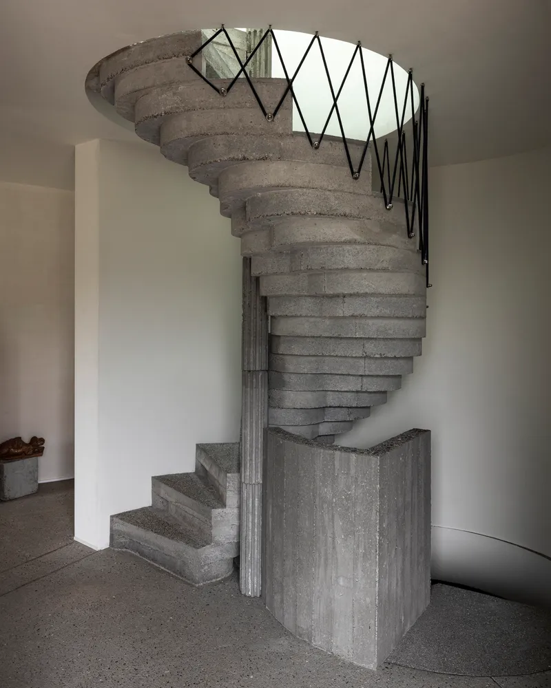 Helical concrete stair with zigzag metal railing and board-formed central column rising through circular opening