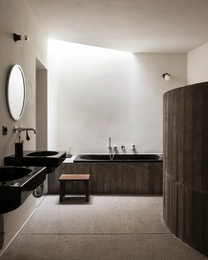 Bathroom with black vessel sinks, board-formed tub surround, and skylight washing white plaster walls