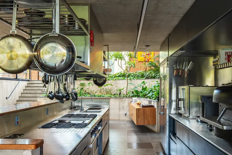 Galley kitchen with stainless steel appliances, exposed concrete ceiling and hanging cookware overlooking a planted terrace