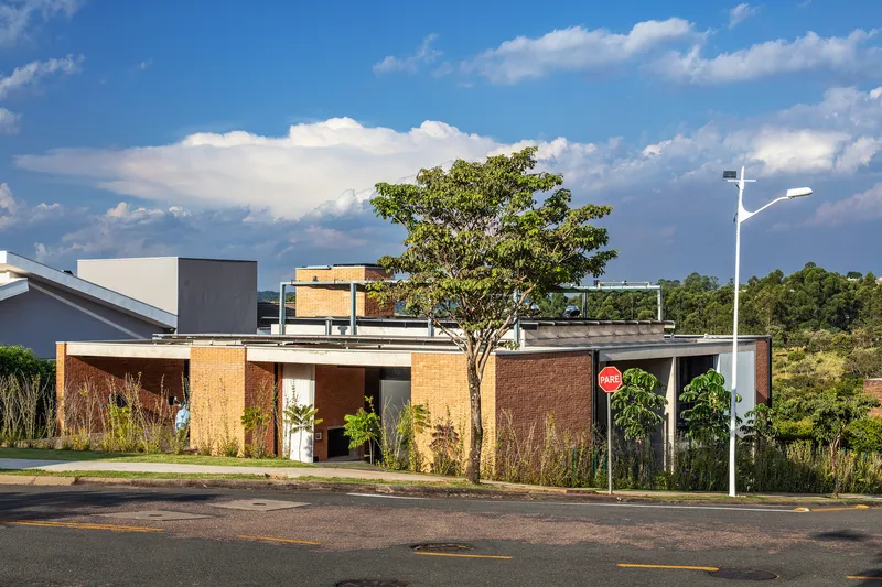 Street view of the low-slung residence with brick walls, planted garden beds and rooftop terrace under scattered clouds