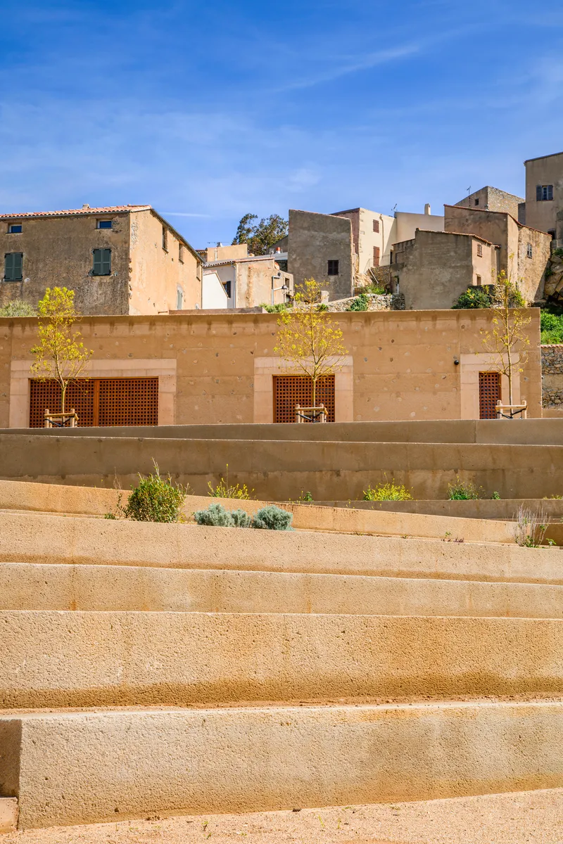 Stepped plaza with young trees and wood-slatted openings in the concrete retaining wall below the old village