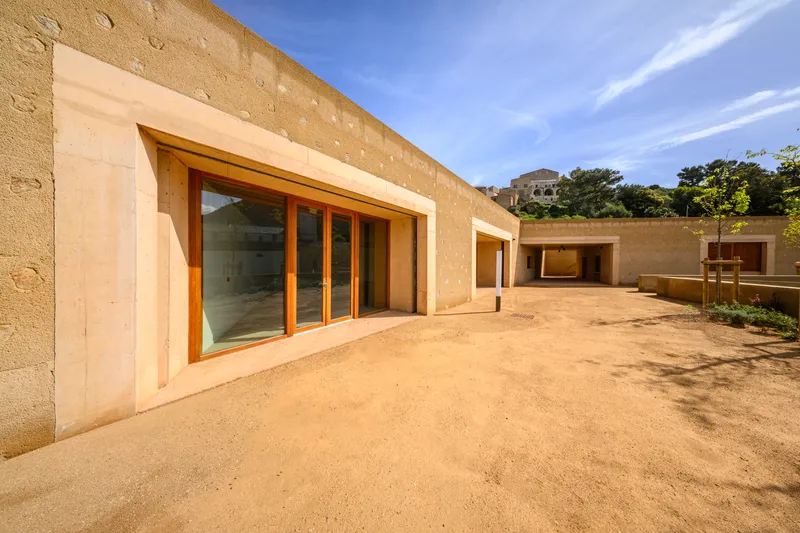 Exterior courtyard with rammed earth walls and timber-framed glazing under a blue afternoon sky