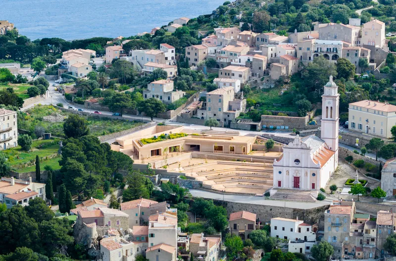 Aerial view of the terraced amphitheater and low-slung volumes nestled among hillside houses and a white church tower