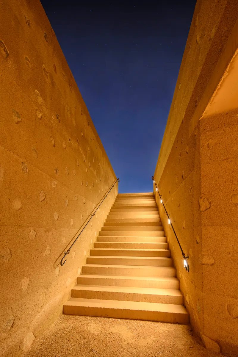 Concrete staircase ascending between rammed earth walls toward a narrow opening framing the night sky
