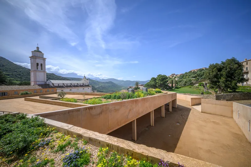 Elevated concrete roof terrace with planted beds alongside a historic church tower and mountains