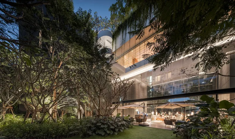 Glass-wrapped facade with cantilevered terraces surrounded by mature trees and tropical planting at dusk