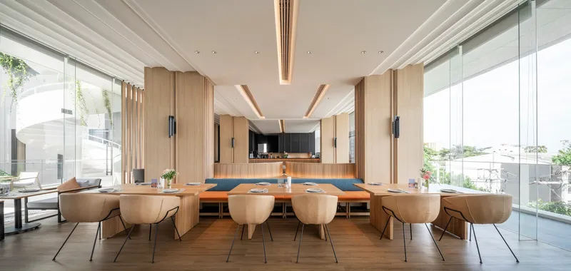 Communal dining space with long timber table, upholstered seating, and vertical wood paneling framing the kitchen beyond
