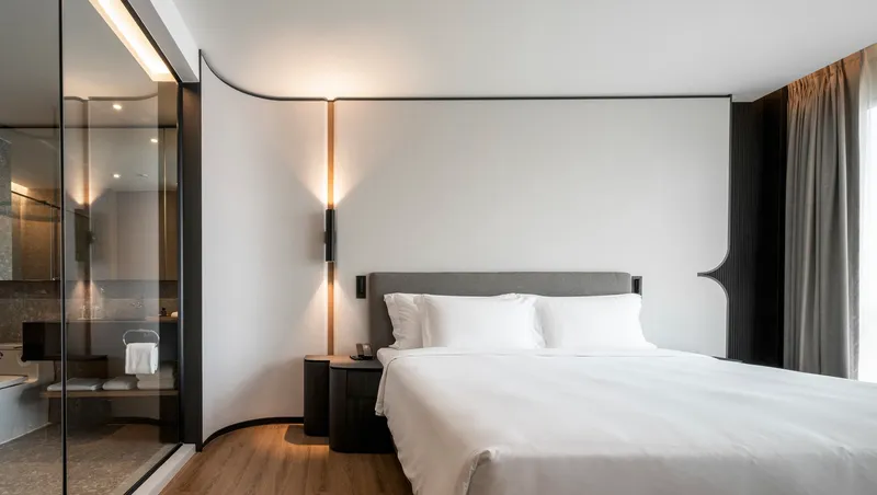 Hotel bedroom with upholstered headboard, bronze wall sconces, and glass-enclosed bathroom at dusk