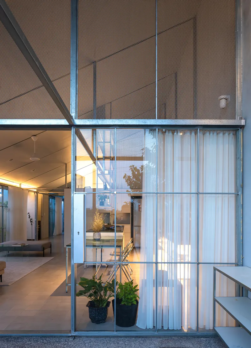 Two-story steel-framed glass partition with white curtains and potted plants dividing interior spaces