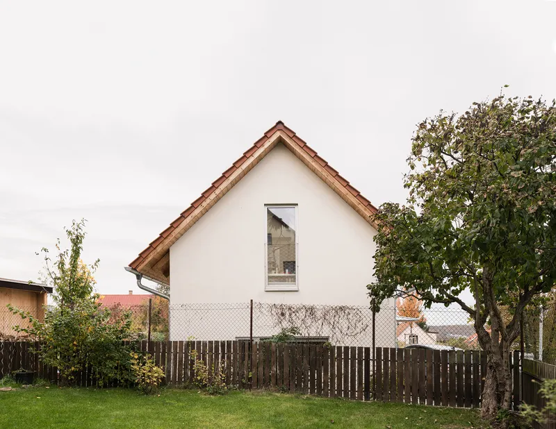White rendered gable facade with single tall window viewed across lawn and weathered timber fence