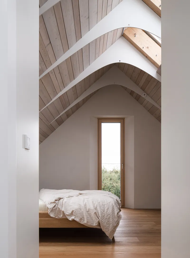 Bedroom with exposed timber rafters, tall narrow window and platform bed under vaulted ceiling