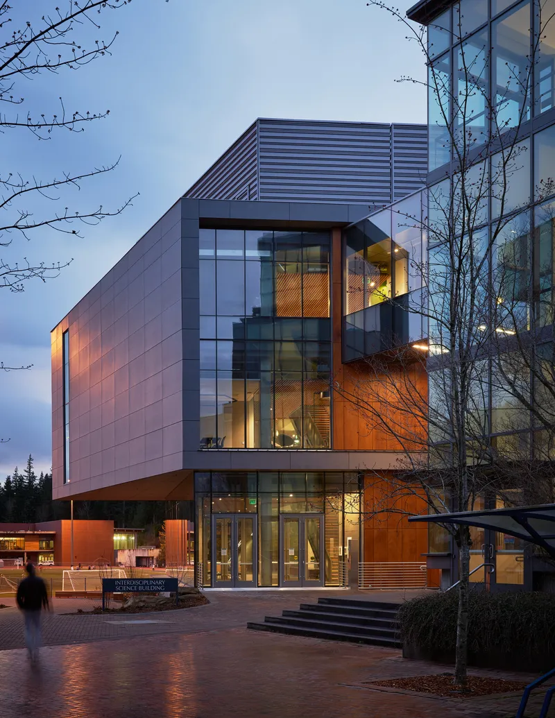 Illuminated entrance with cantilevered volumes and weathered steel panels at dusk with bare tree branches