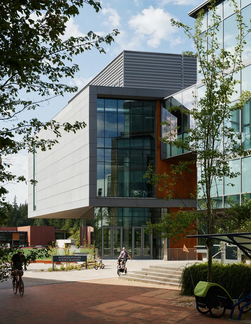 Entrance facade with cantilevered volumes and weathered steel panels framed by trees and cyclists on the plaza