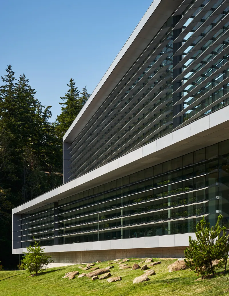Layered horizontal louver system wrapping the building facade above a landscaped slope with scattered boulders