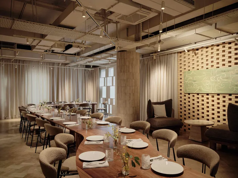 Long dining tables set with white plates beneath exposed ceiling ducts and sheer curtains filtering daylight