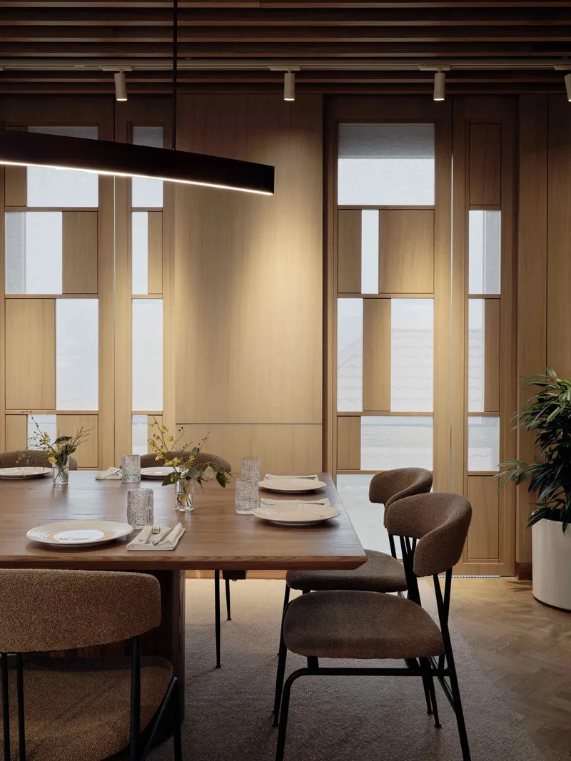 Dining table beneath a linear black pendant with timber framed glass partitions in soft daylight