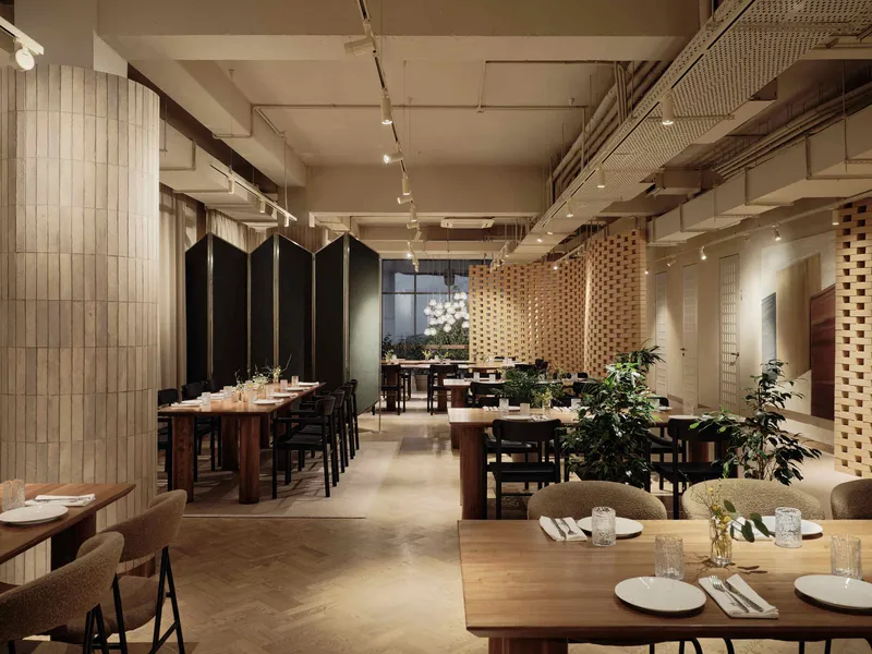 Dining hall showing woven timber partition wall, vertical black folding screens, and potted plants between table groupings