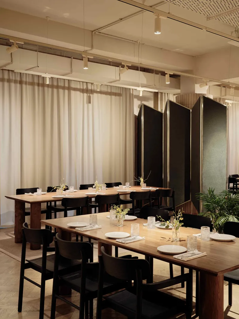 Two timber dining tables with black chairs separated by black folding screens and sheer curtains