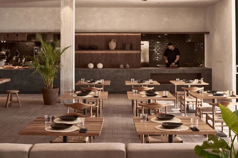 Restaurant dining room with concrete columns, timber tables and a chef working at the open kitchen