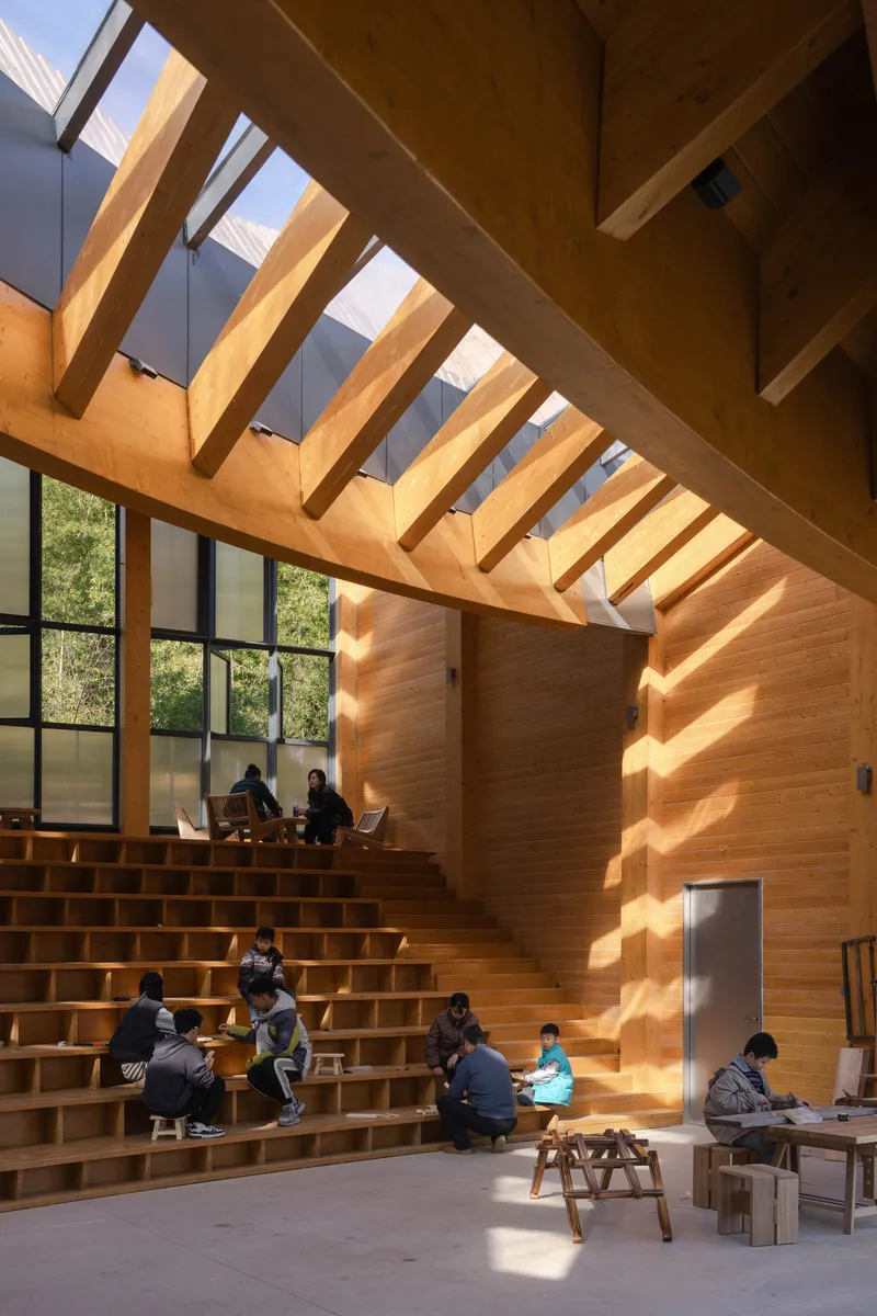 Stepped timber seating with visitors gathered below exposed wood beams and tall glazed walls in afternoon light