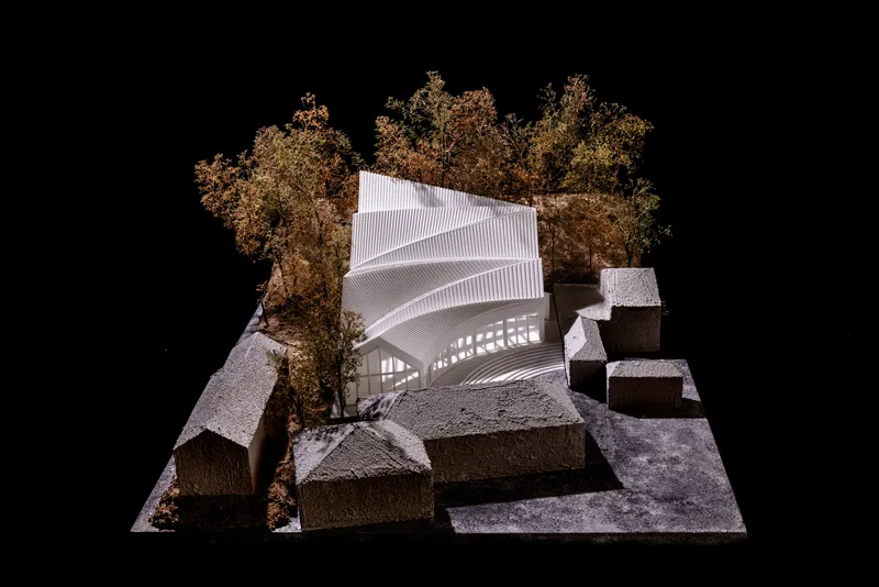 Aerial view of physical model showing ribbed white roof structure nestled among autumn trees and grey block forms