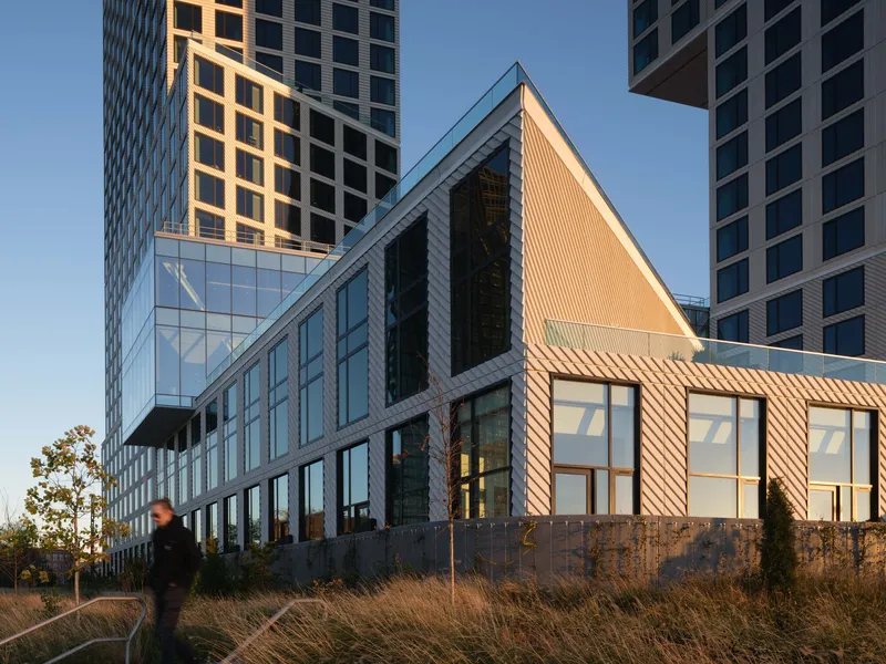 Striped pavilion with peaked roof and glazed facade beside high-rise towers and a person walking through native grasses