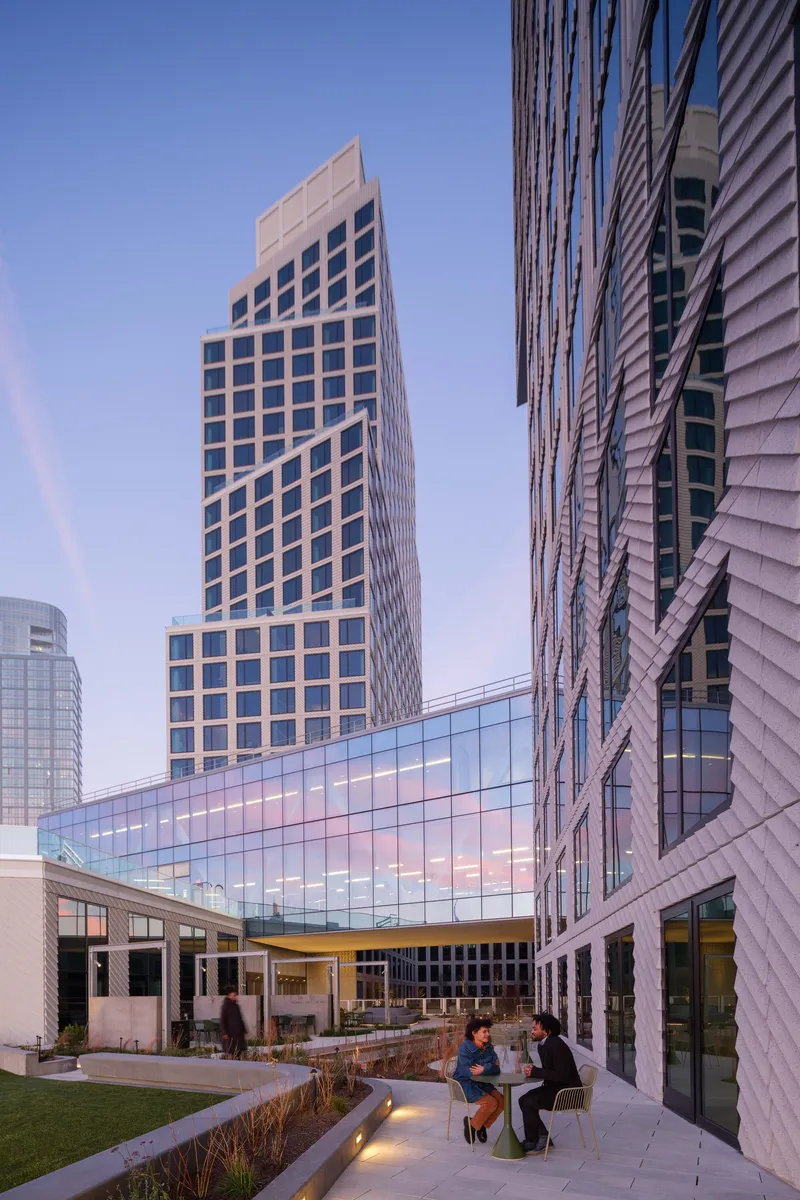 Rooftop terrace with two people seated at dusk beneath a glass bridge connecting residential towers
