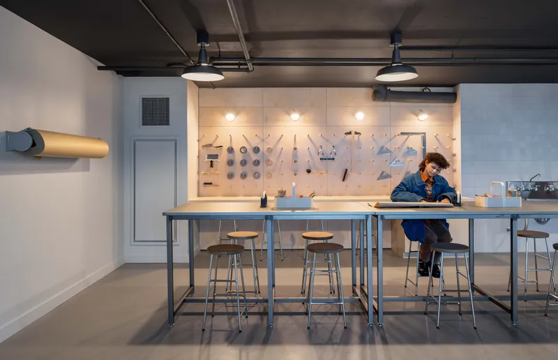Workshop space with tool wall and high metal tables where a person sits under pendant lights