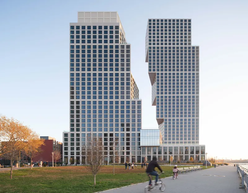 Two residential towers with gridded facades beside a waterfront promenade with cyclists and autumn trees