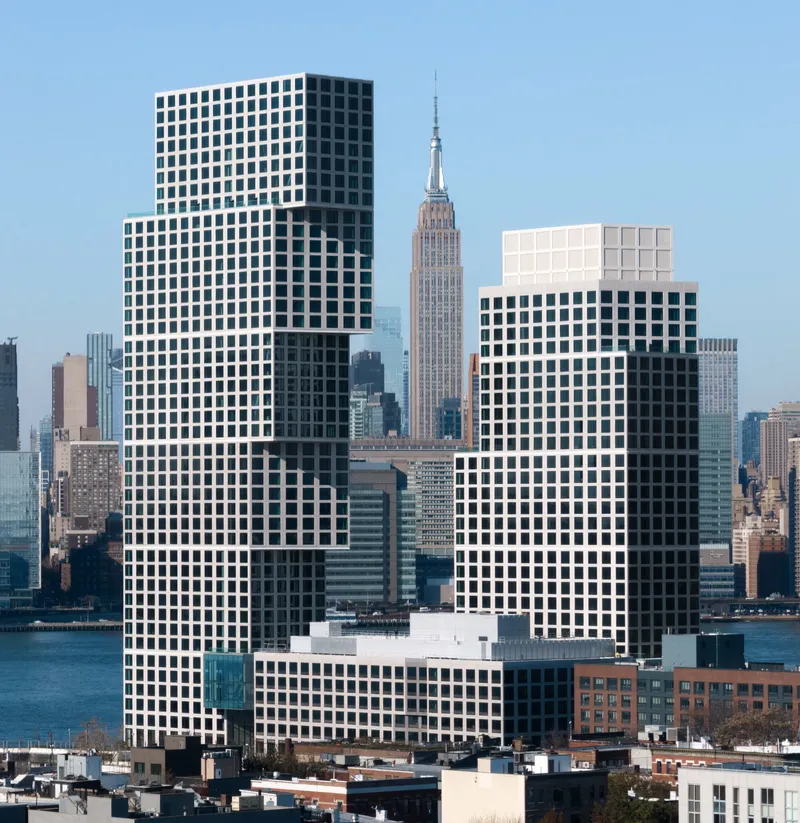 Three stacked volumes with punched window grids rising above the waterfront neighborhood under clear sky