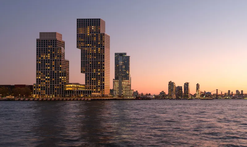 Waterfront view of three staggered residential towers with illuminated windows at sunset