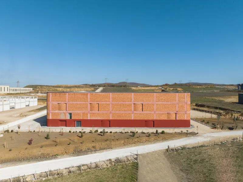 Long facade of terracotta brick panels over red base with newly planted saplings along access road