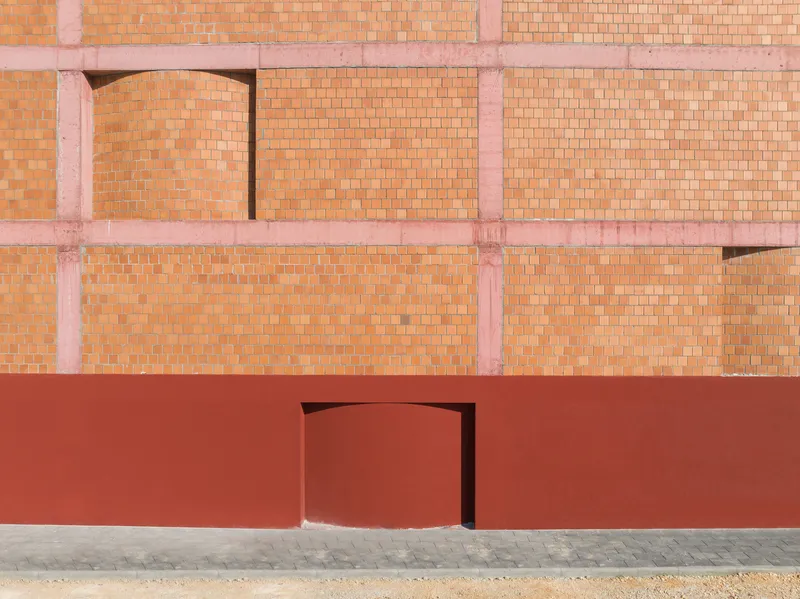 Curved red entry door set into red plinth below terracotta brick facade with arched openings