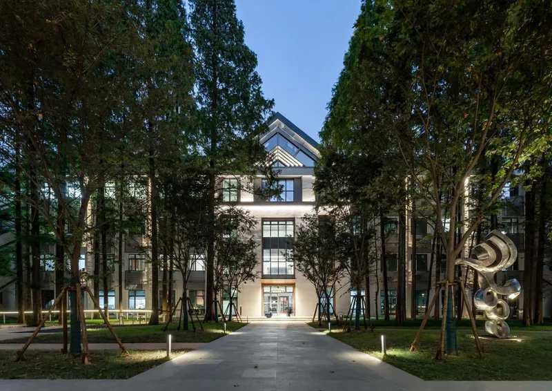 Front facade illuminated at dusk, framed by rows of tall trees along a paved walkway