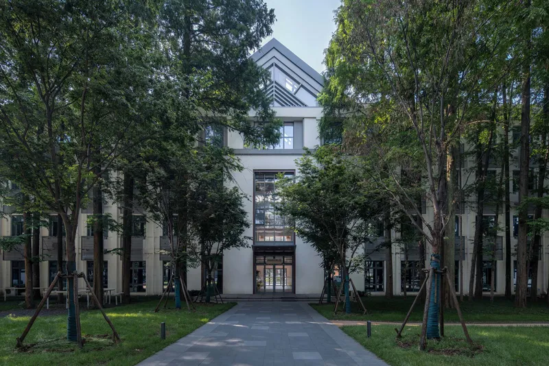 Tree-lined pedestrian path leading to the glazed entrance volume under a pale blue sky