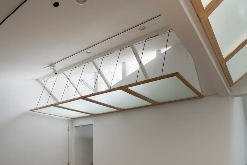 Diagonal lattice glazing system above a suspended timber-framed panel in a white interior