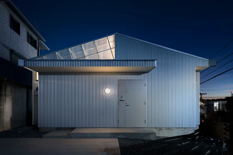 Corrugated metal facade with illuminated sawtooth roof and entry door at twilight