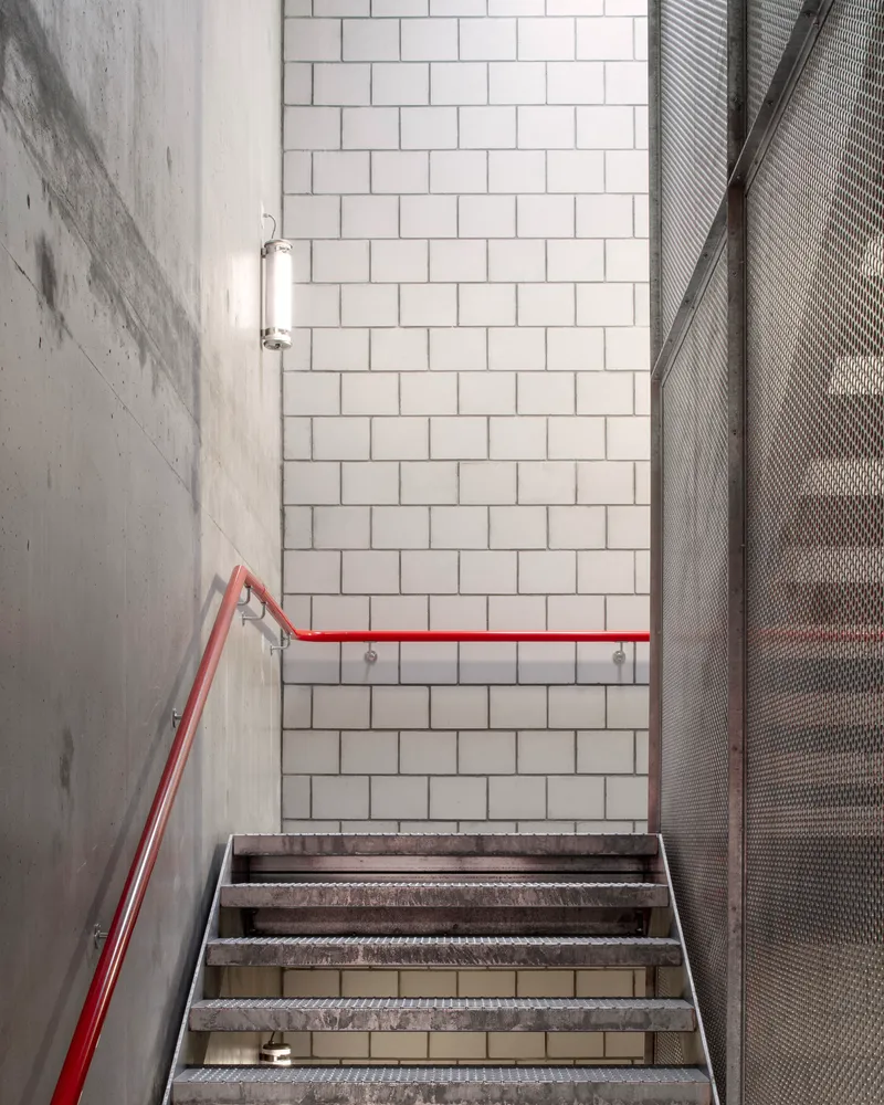 Concrete stairwell with red handrail, white tile wall and perforated metal screen panel