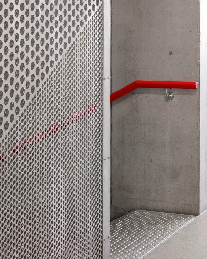 Perforated metal screen wall meeting a concrete wall with a red painted handrail at the landing