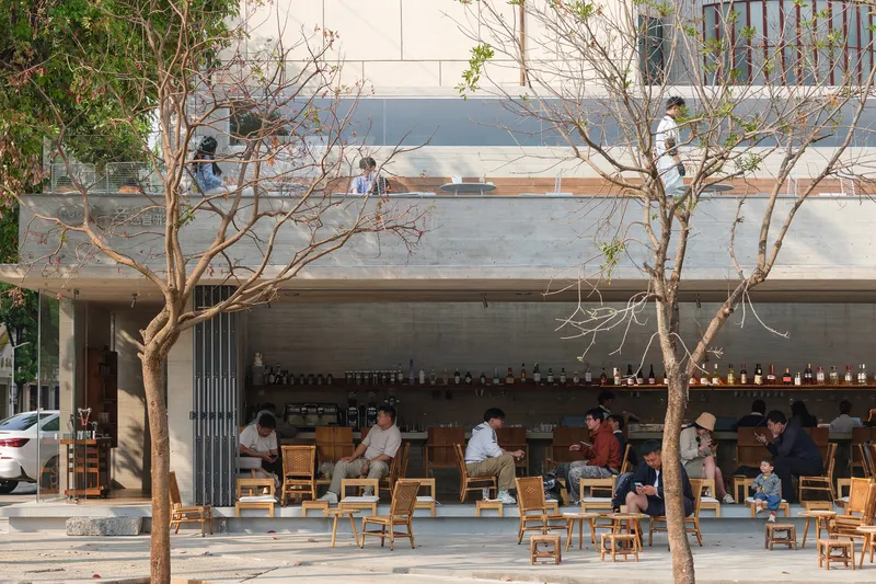 Cafe terrace with wooden seating under concrete canopy framed by bare branched trees