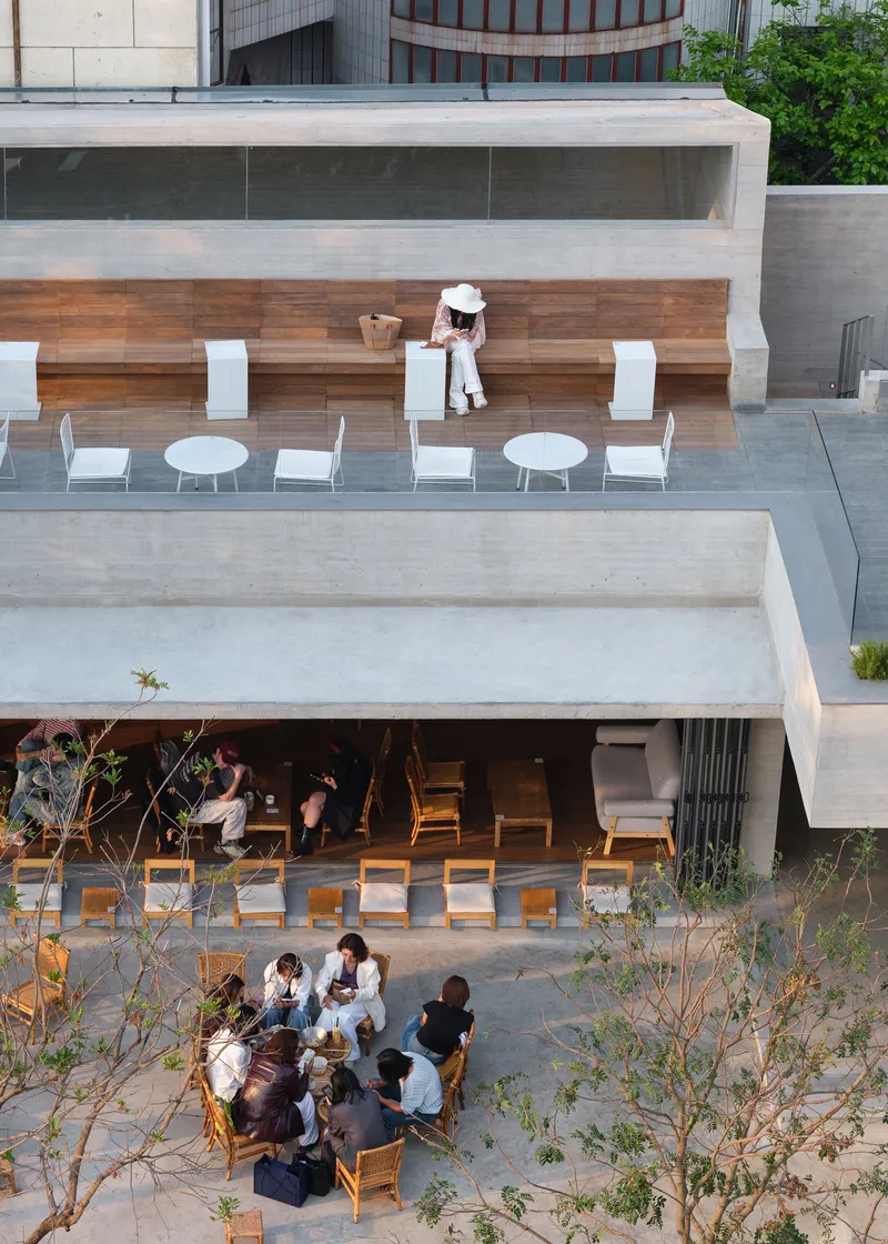 Overhead view of stacked concrete terraces with timber benches and white cafe seating among young trees