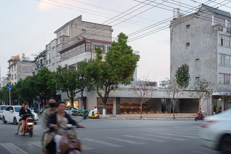 Street corner facade with recessed glazing and mature trees as motorbikes pass at dusk