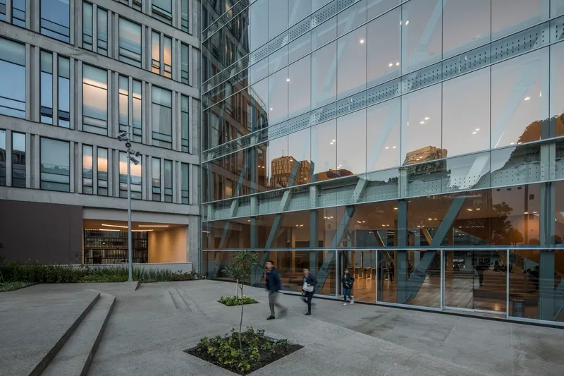 Evening courtyard view showing the angled steel braces behind the glass ground floor facade with people passing