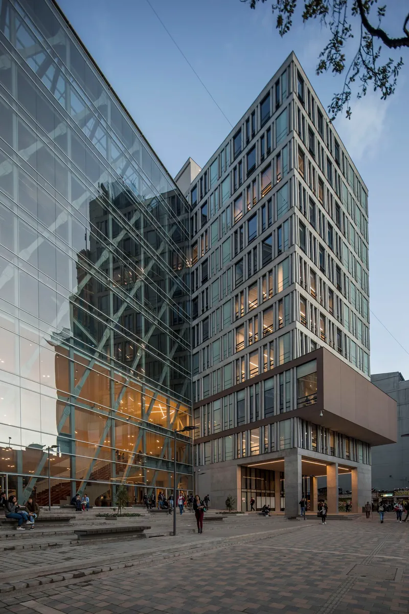 Glazed tower facade and cable-supported glass atrium above a paved public plaza at dusk
