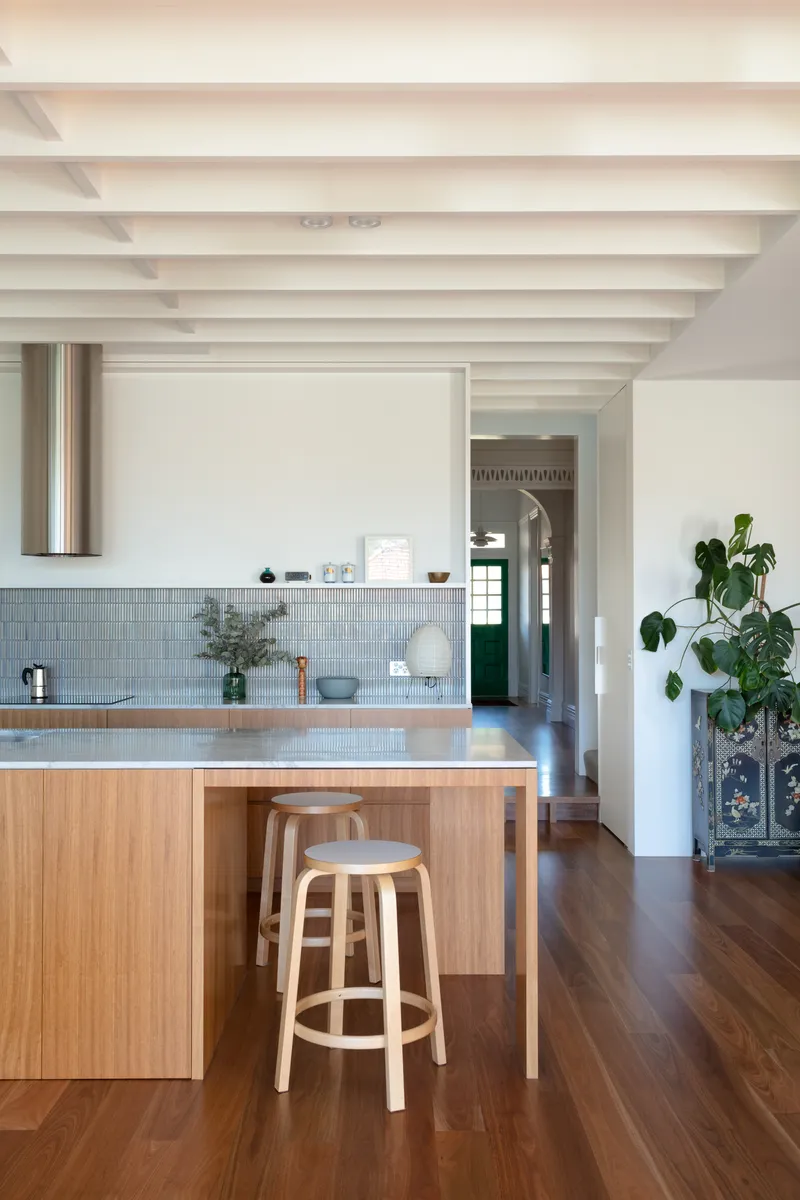 Kitchen island with pale timber cabinetry and grey tile countertop under painted beam ceiling